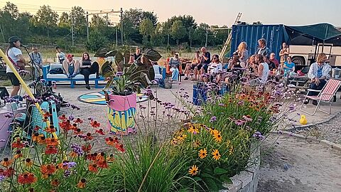 Mensen zitten op stoelen en kijken naar een optreden van een zanger met gitaar. Op de voorgrond staat een grote bak met planten.