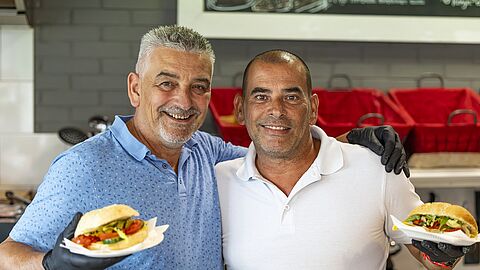 2 mannen naast elkaar achter de toonbank van een broodjeszaak. Beiden hebben ze een belegd broodje in hun hand. 